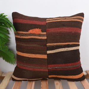 Peut inclure: Un coussin carré décoratif avec un motif patchwork. Le coussin présente des rayures horizontales dans les tons de marron, d'orange et de rouge. Le coussin est posé sur une surface à lattes en bois, avec une plante verte en arrière-plan.