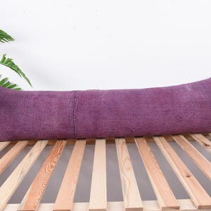 May include: A long, rectangular purple pillow with a textured surface. The pillow is placed on a wooden slatted frame. A houseplant in a wooden planter is visible in the background.