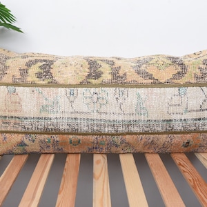 May include: Rectangular decorative pillow with a vintage, distressed look. The pillow features a patchwork design with a mix of colors including beige, brown, and blue. The pillow is placed on a wooden bed frame.