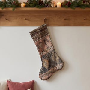 May include: A Christmas stocking featuring a faded, distressed rug-like pattern in muted tones of dusty pink, charcoal grey, and beige. The stocking has a textured, woven appearance with a hanging loop at the top, suspended from a wooden peg.