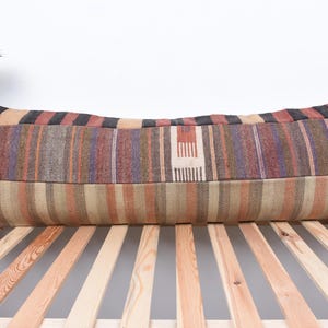 Peut inclure: Un long coussin rectangulaire avec un motif rayé dans les tons de brun, beige, rouge et violet. Le coussin est fait de tissu tissé et a une surface légèrement texturée.