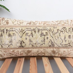 Puede incluir: Cojín rectangular beige con un diseño detallado en tonos marrón y verde. Se encuentra sobre una base de listones de madera, con un fondo blanco y una hoja verde en la esquina superior izquierda.