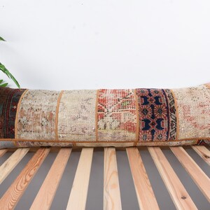 Puede incluir: Una almohada larga y rectangular hecha de tela de patchwork. La almohada está hecha de diferentes piezas de tela en tonos beige, marrón, rojo y azul. La almohada está sobre una superficie de madera.
