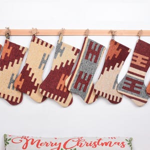 Può includere: Otto calze di Natale con motivi geometrici nei toni del rosso, beige e grigio, appese a un'asta di legno. Le calze sono realizzate in tessuto intrecciato e sono esposte su uno sfondo bianco. Ogni calza ha un design unico.