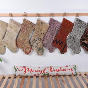May include: Eight colorful Christmas stockings made from repurposed rugs hang on a wooden rod. The stockings have various patterns and colors, including red, blue, green, and brown. A white pillow with the text "Merry Christmas" is on a wooden bench.