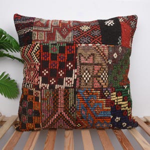 May include: A square patchwork pillow with a variety of geometric patterns in shades of red, black, green, and orange. The pillow is placed on a wooden slatted surface, with a green plant in the background.