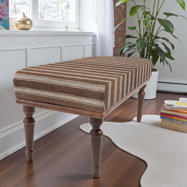 May include: A rectangular bench with a striped brown and beige upholstered top. The bench has four turned wooden legs. The legs are a light brown colour with darker brown accents. The bench is placed on a hardwood floor.