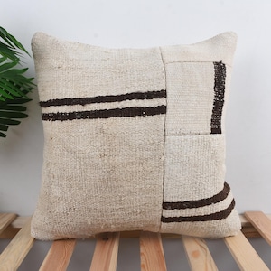 Peut inclure: Coussin carré crème avec des rayures marron foncé et des patchs géométriques. Le coussin est fait d'un tissu texturé et repose sur une surface à lattes en bois. Décoration d'intérieur.