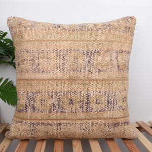 Peut inclure: Coussin carré beige et marron avec un motif géométrique. Le coussin présente des rayures horizontales et un aspect texturé. Le coussin est posé sur une surface en bois, avec une plante verte en arrière-plan.