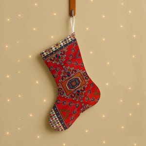 Könnte beinhalten: Ein roter Weihnachtsstrumpf mit einem detaillierten Muster, der an einem Holzhaken hängt. Das Muster enthält geometrische Formen und eine weiße Schlaufe. Der Hintergrund ist neutralfarben mit kleinen, funkelnden Lichtern.