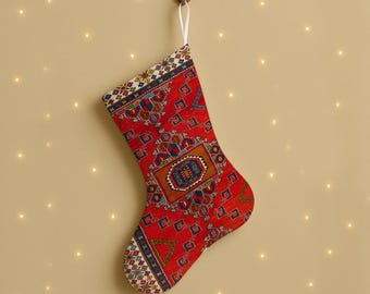 Puede incluir: Un calcetín navideño rojo con un diseño detallado, colgado de un gancho de madera. El diseño incluye formas geométricas y un lazo blanco. El fondo es de color neutro con pequeñas luces parpadeantes.