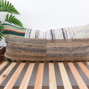 Op de afbeelding: Een lang, rechthoekig kussen met een patchwork-ontwerp gemaakt van verschillende stoffen. Het kussen is gemaakt van verschillende kleuren, waaronder bruin, beige, wit en zwart. Het kussen ligt op een houten lattenbodem.