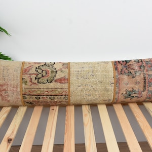 May include: A long, rectangular cushion with a patchwork design. The cushion is made of beige, pink, and blue fabric with intricate patterns. The cushion is lying on a wooden slatted bed frame.