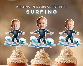 Puede incluir: Cupcakes con adornos para cupcakes con temática de surf. Cada adorno presenta un bebé surfista de dibujos animados con un traje de neopreno negro sobre una tabla de surf azul y roja. Los cupcakes tienen glaseado marrón claro. El texto en la imagen dice: "PERSONALIZED CUPCAKE TOPPERS SURFING".