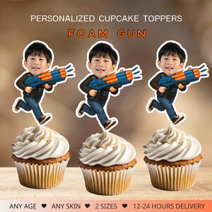 May include: Cupcake toppers featuring a cartoon boy with a blue and orange foam gun. The toppers are placed on top of cupcakes with white frosting. Text on the image reads "PERSONALIZED CUPCAKE TOPPERS" and "FOAM GUN".
