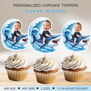 Może przedstawiać: Trzy babeczki zwieńczone spersonalizowanymi topperami z rekinami. Każdy topper przedstawia kreskówkowe dziecko jadące na rekinie na tle niebieskiej fali. Babeczki mają białe lukier i są w brązowych papierowych foremkach. U góry widnieje napis "PERSONALIZED CUPCAKE TOPPERS SHARK RIDING."