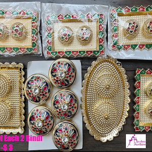 May include: Assortment of gold-colored trays with decorative designs, some with red and green accents. The trays are filled with small, round, gold-colored containers with colorful floral designs and a single rhinestone in the center. The text "Price of Each 2 Bindi box US - $3" is visible at the bottom of the image.
