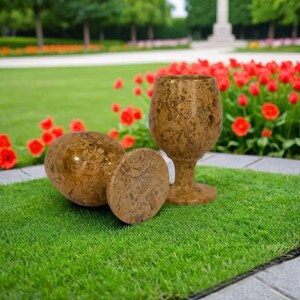 May include: A set of decorative items, including a goblet, and two round objects, all with a mottled brown pattern. The objects are arranged on a patch of green artificial grass, with a blurred background of red flowers and a park.