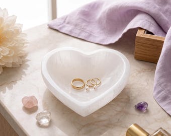 May include: A white heart-shaped bowl made of polished stone with a smooth, translucent finish. Inside the bowl rests a plain gold band and a pair of small gold hoop earrings featuring tiny white pearl accents, creating a simple jewellery display.