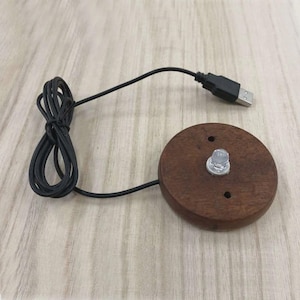 Op de afbeelding: Een ronde, donkerbruine houten basis met een helder LED-licht en een zwarte USB-kabel. De basis heeft twee kleine gaten en een zilverkleurige moer om het licht te bevestigen. De USB-kabel is opgerold en verbonden met de basis.