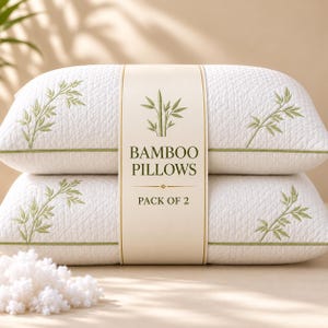 May include: A pack of two white pillows with a quilted texture and green embroidered bamboo leaf patterns. A central paper band features a bamboo illustration and the text BAMBOO PILLOWS PACK OF 2. A small pile of white pillow filling sits in the foreground.