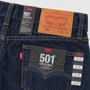May include: Dark blue denim jeans featuring a tan leather patch with Levi Strauss & Co. branding, a red tab on the back pocket, and multiple black paper tags displaying 501 Original, 100% cotton, and size W 34 L 40 specifications.