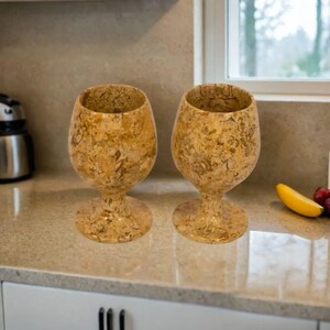 May include: Two decorative wine glasses with a speckled, beige and brown pattern. The glasses are made of a stone-like material and have a wide bowl and a stemmed base. They are displayed on a light-colored countertop.