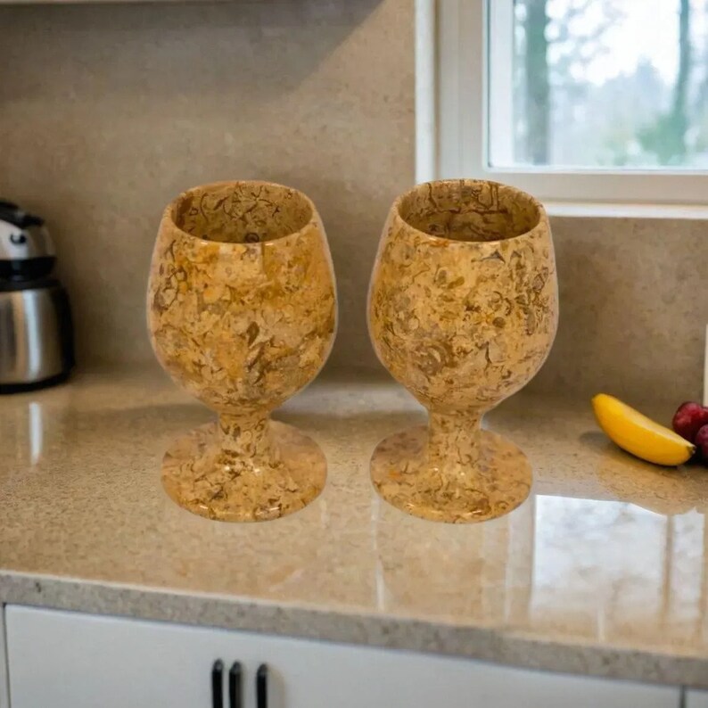 May include: Two decorative wine glasses with a speckled, beige and brown pattern. The glasses are made of a stone-like material and have a wide bowl and a stemmed base. They are displayed on a light-colored countertop.