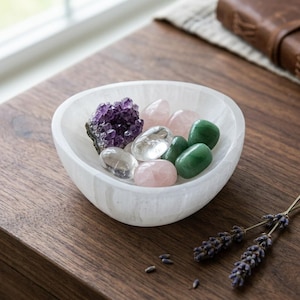 May include: A white, translucent, bowl-shaped selenite dish containing a collection of polished gemstones, including a dark purple amethyst cluster, clear quartz, pale pink rose quartz, and smooth green aventurine stones.