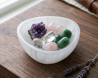 May include: A white, translucent, bowl-shaped selenite dish containing a collection of polished gemstones, including a dark purple amethyst cluster, clear quartz, pale pink rose quartz, and smooth green aventurine stones.