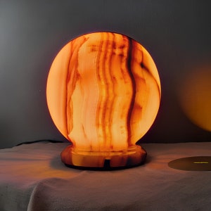 Peut inclure: Une lampe ronde en onyx orange et marron avec une lueur blanche. La lampe est posée sur une surface blanche.