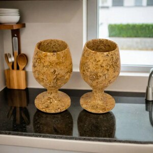 May include: Two decorative wine glasses with a speckled, tan and brown pattern. The glasses have a rounded bowl shape and sit on a short stem with a circular base. They are displayed on a black countertop.