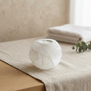 May include: A spherical white candle holder with a faceted, geometric surface texture. The object features a circular opening at the top designed to hold a small candle, showcasing a translucent, layered appearance with subtle white and grey tones.