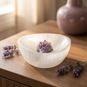 May include: A white, translucent, bowl-shaped selenite dish containing a small, jagged purple amethyst crystal cluster. The bowl has a smooth, rounded rim and a fibrous, natural texture, contrasting with the sharp, crystalline points of the amethyst.