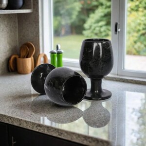 May include: Three black marble wine glasses on a countertop. One glass is upright, while the other two are lying on their sides. The glasses have a dark, veined pattern and a glossy finish. The countertop is light gray.