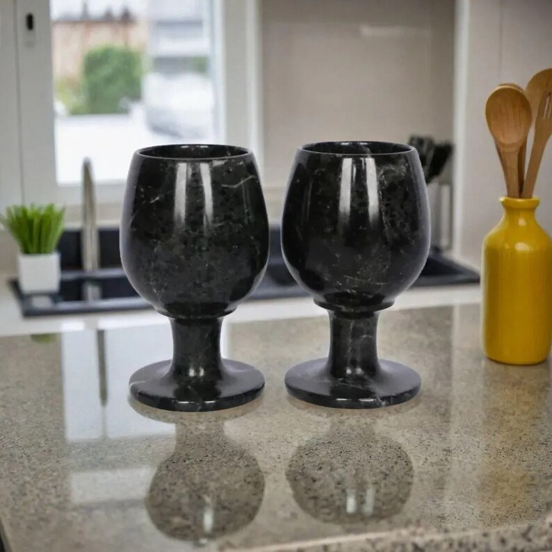 May include: Two black marble wine glasses with a speckled pattern. The glasses have a rounded bowl shape, a stem, and a circular base. They are displayed on a light-colored countertop, reflecting their shape. The kitchen setting includes a yellow vase and a small plant.
