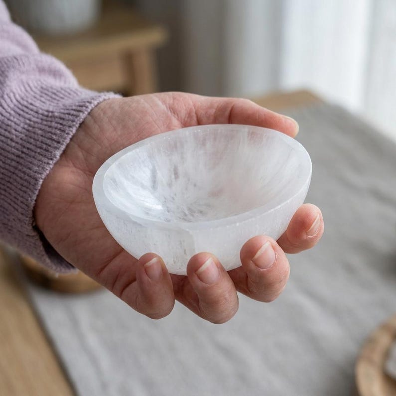May include: A small, round, translucent white selenite bowl with a smooth, polished interior and a natural, fibrous crystalline texture. The bowl features a shallow basin and a rounded rim, showcasing the mineral's characteristic pearly luster.
