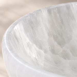May include: A white selenite bowl with a smooth, polished exterior rim and a textured, crystalline interior surface. The translucent stone features natural vertical striations and a fibrous, layered appearance throughout the curved structure.