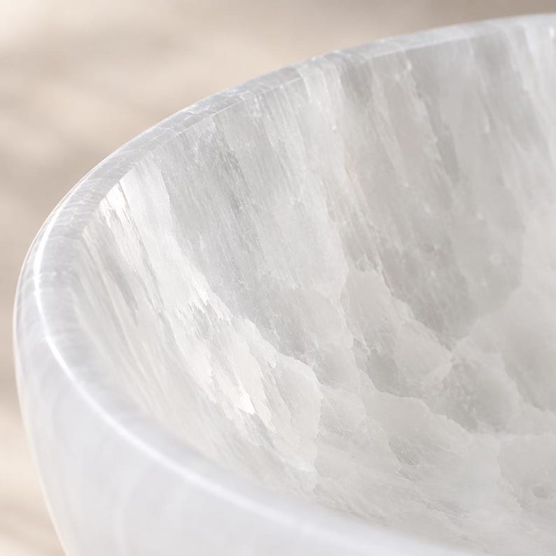 May include: A white selenite bowl with a smooth, polished exterior rim and a textured, crystalline interior surface. The translucent stone features natural vertical striations and a fibrous, layered appearance throughout the curved structure.