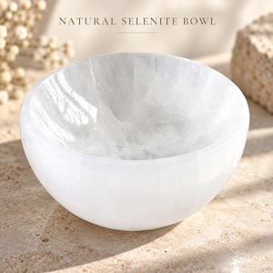 May include: A round, white, translucent bowl carved from natural selenite with a smooth, polished exterior and a textured, crystalline interior. The text NATURAL SELENITE BOWL is printed in dark lettering above the object.