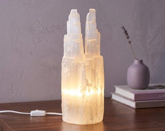 May include: A cylindrical selenite crystal lamp with a jagged, tower-like top edge. The translucent white stone emits a warm, internal glow from the base. A white electrical cord with a small rectangular toggle switch extends from the side.
