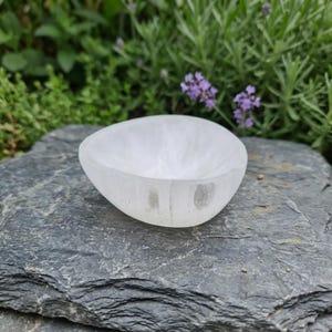 May include: A small, smooth, white selenite bowl with a rounded, organic shape. The bowl has a translucent, crystalline appearance with subtle variations in opacity and a polished finish, showcasing a simple, hollowed-out center design.