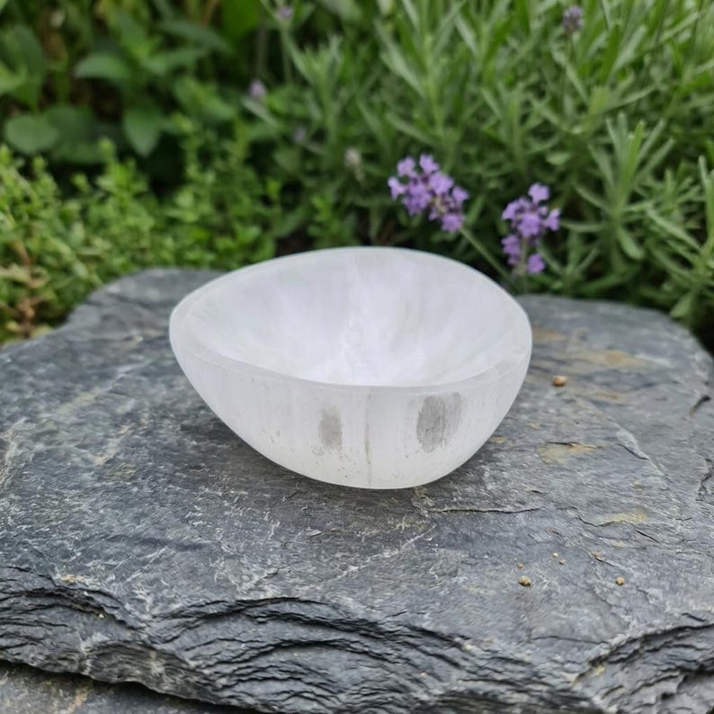 May include: A small, smooth, white selenite bowl with a rounded, organic shape. The bowl has a translucent, crystalline appearance with subtle variations in opacity and a polished finish, showcasing a simple, hollowed-out center design.