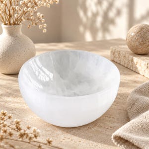 May include: A polished white selenite bowl with a smooth, rounded exterior and a translucent, crystalline interior texture. The bowl features a deep, curved basin and a solid, opaque white base, showcasing natural mineral veining throughout.