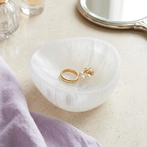 May include: A small, white, translucent, organic-shaped bowl with a marbled texture. Inside the bowl sits a plain gold band ring and a pair of gold stud earrings featuring small white pearls. The bowl is designed to hold small jewelry items.