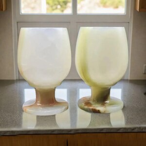 May include: Two elegant, translucent wine glasses with a rounded bowl and a short stem. The glasses are made of a light-colored stone, with variations of white, cream, and green. They sit on a reflective surface, suggesting a kitchen or dining setting.