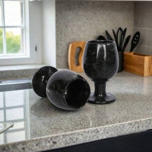 May include: Three black marble wine glasses on a countertop. Two glasses are lying on their sides, while the third stands upright. The glasses have a dark, veined appearance, and the countertop is a speckled gray.