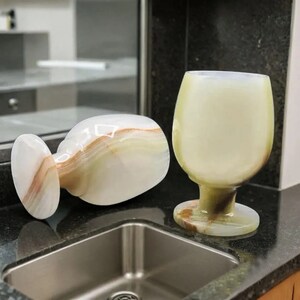 May include: Two onyx wine glasses. One is upright, the other is lying on its side. The glasses have a creamy white and brown color scheme. The stem and base of the glasses are a darker shade of brown.