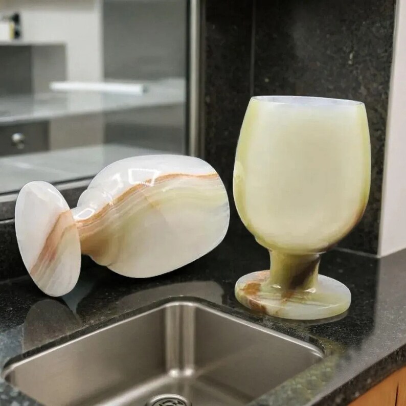 May include: Two onyx wine glasses. One is upright, the other is lying on its side. The glasses have a creamy white and brown color scheme. The stem and base of the glasses are a darker shade of brown.