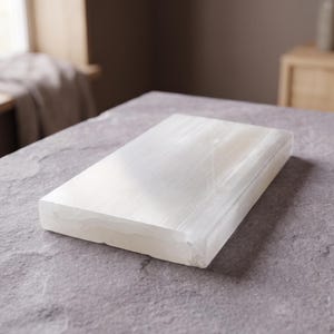 May include: A rectangular slab of white selenite crystal with a smooth, polished top surface and a fibrous, translucent appearance. The stone features natural linear striations and a glossy finish, showcasing its crystalline structure and white color.